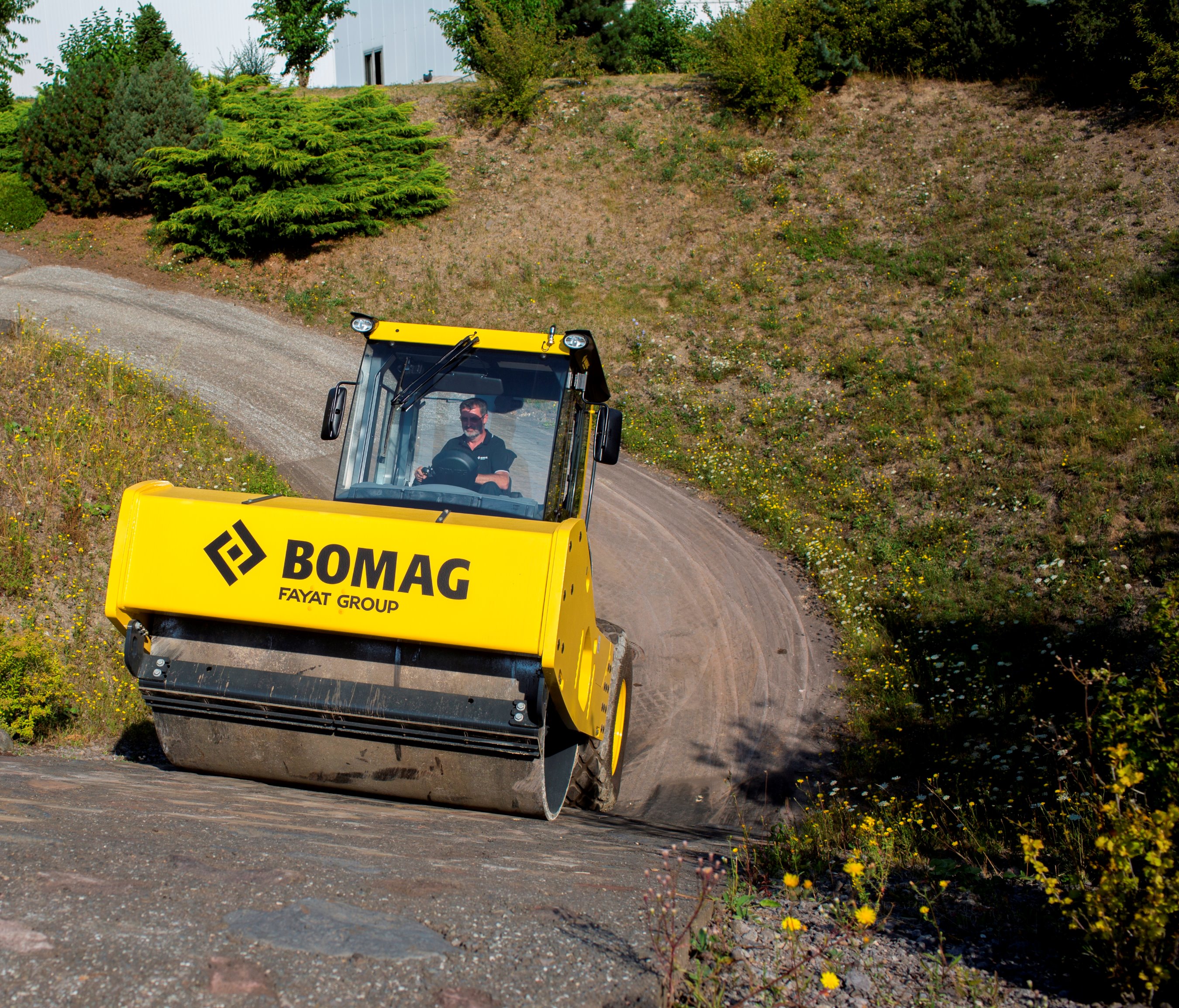 Yellow BOMAG road roller compacting dirt on a curved rural road