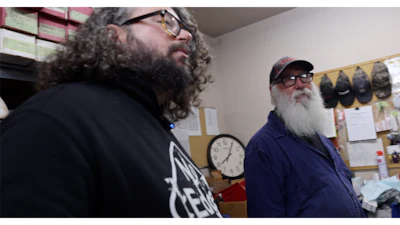 Two men with long beards and glasses standing in an office with a wall clock