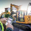 Construction worker holding smartphone monitoring yellow CAT excavator at job site with buildings in background