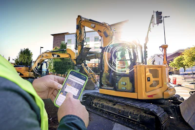 Construction worker holding smartphone monitoring yellow CAT excavator at job site with buildings in background