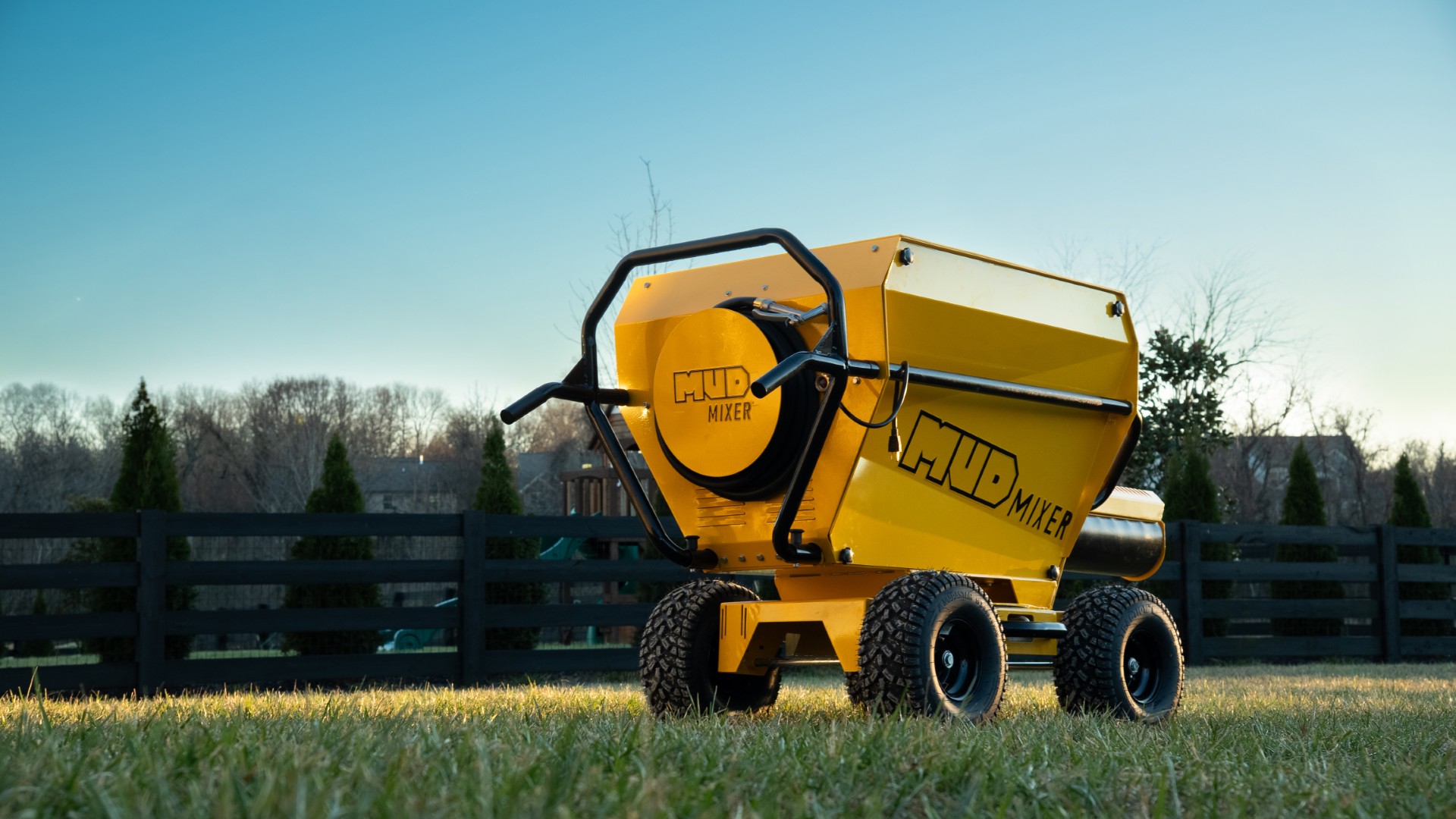 Yellow and black Oshkosh concrete mixer truck parked in a field with wooden fence and trees in background