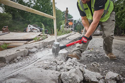 Construction worker using red jackhammer to break concrete on job site with heavy machinery in background
