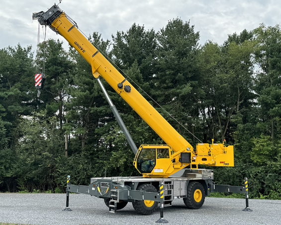 Yellow mobile crane extended with outriggers deployed on gravel lot near pine trees