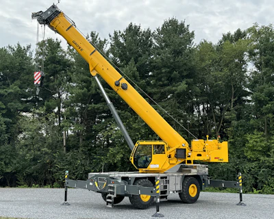 Yellow mobile crane extended with outriggers deployed on gravel lot near pine trees