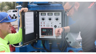 Technicians inspecting blue industrial control panel with multiple gauges and switches
