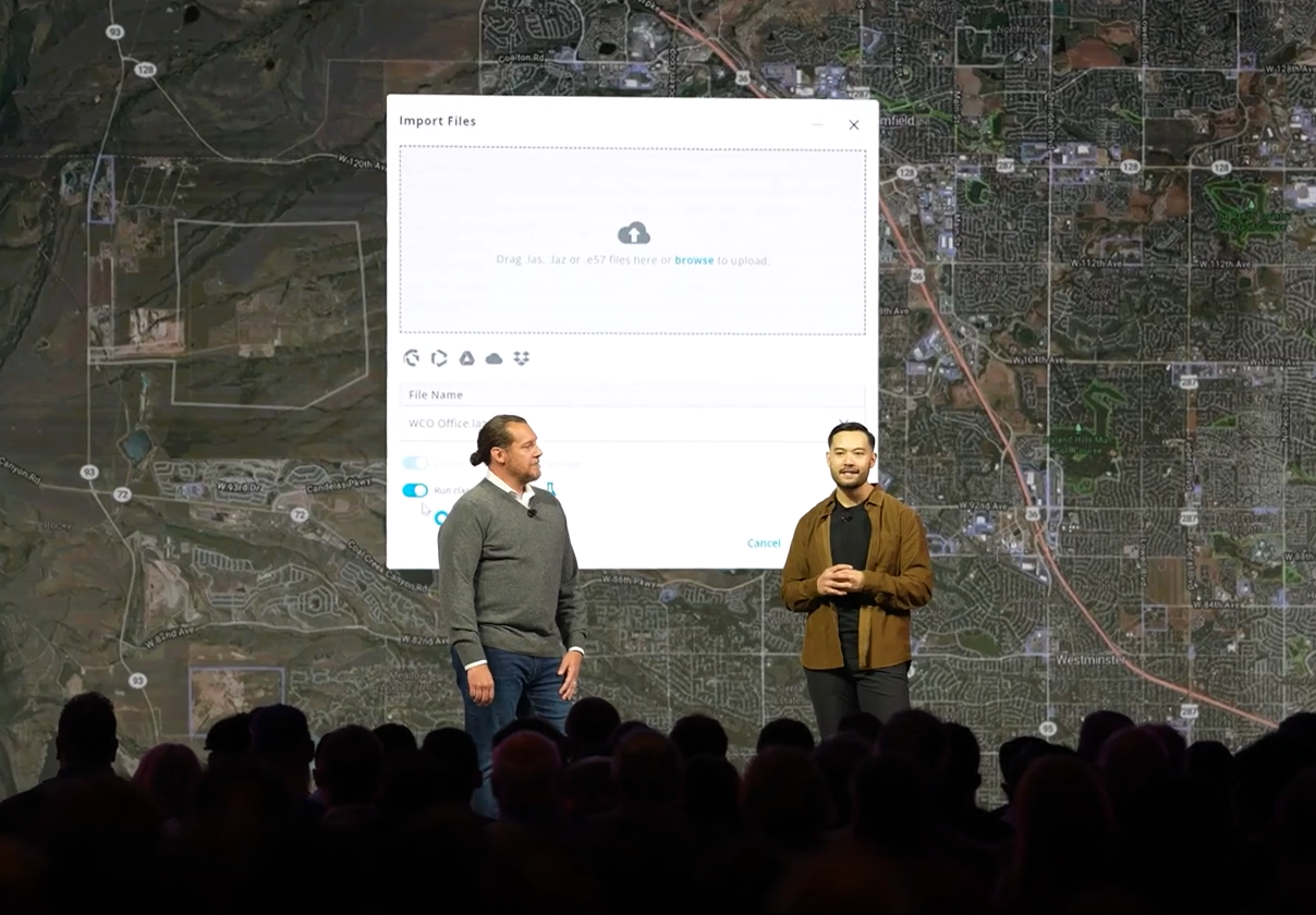 Two presenters on stage discussing mapping software with aerial city map backdrop