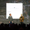 Two presenters on stage discussing mapping software with aerial city map backdrop