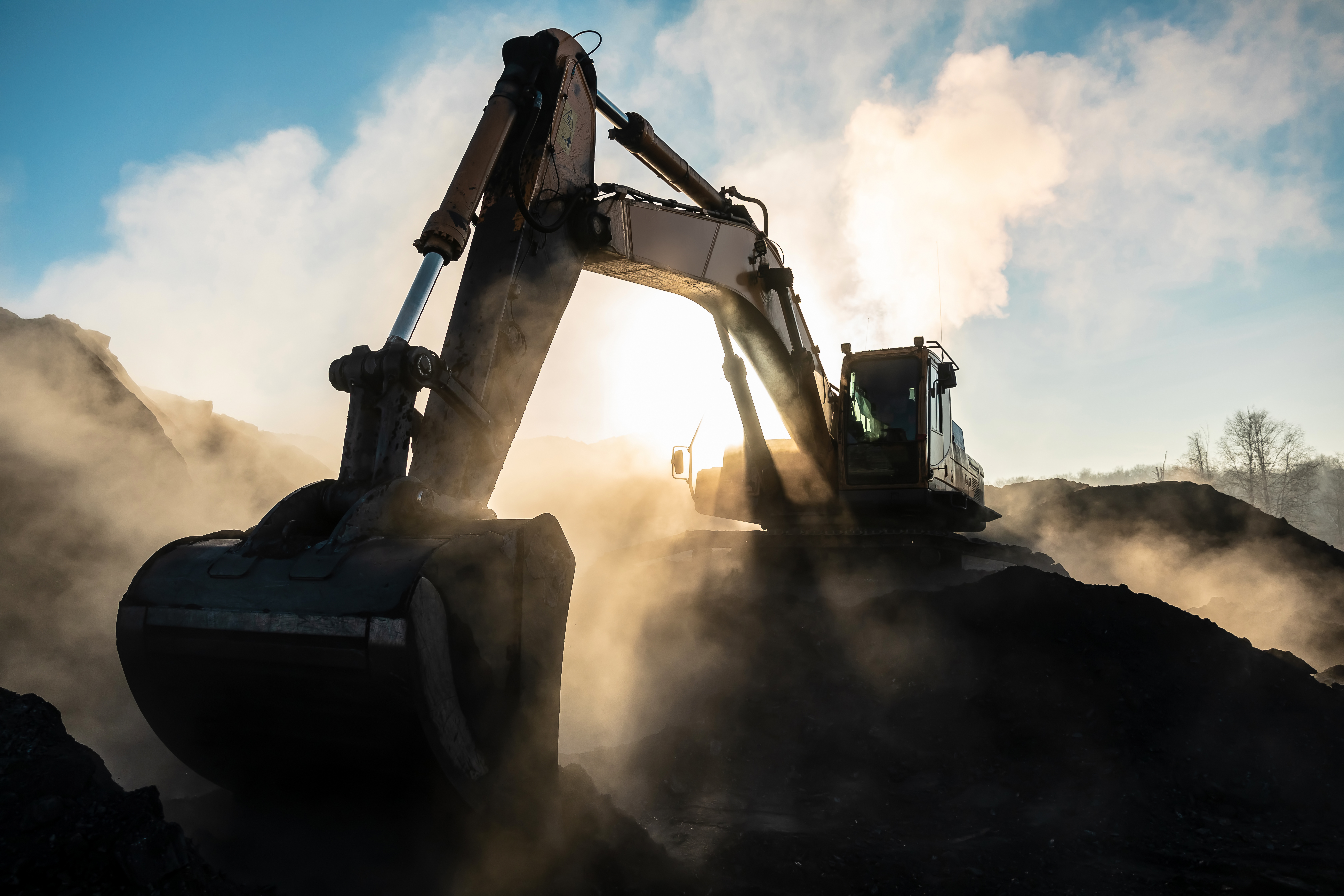 Excavator operating on dusty construction site with blue sky and clouds in background