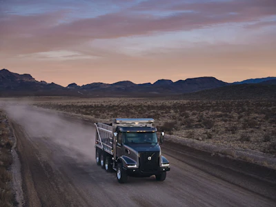 Black Volvo dump truck driving on dusty desert road at sunset with mountains in background