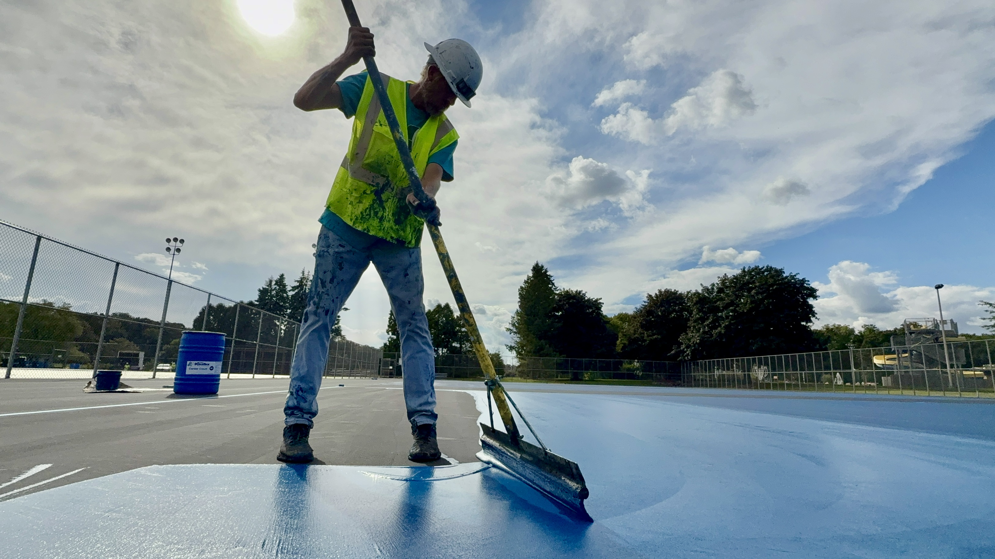 Construction worker in safety vest sweeping blue surface with push broom outdoors