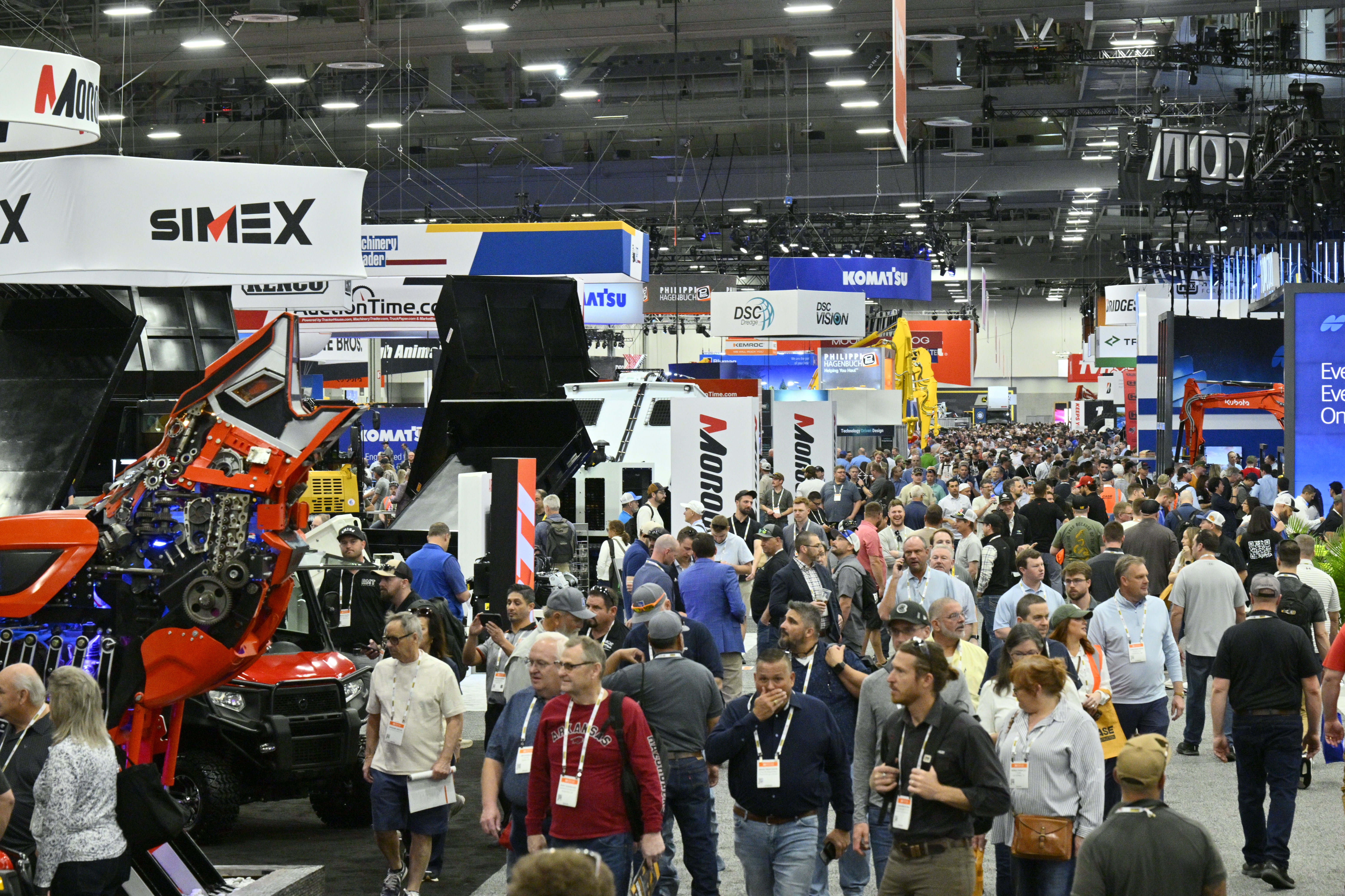 Crowded industrial trade show floor with heavy machinery displays, vendor booths, and attendees wearing identification badges.