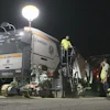 Wirtgen asphalt paver machine operating at night with workers in safety gear and illuminated work lights