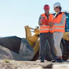 Two construction workers in safety vests review plans at a job site with heavy machinery