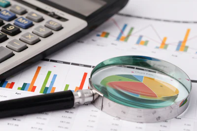 Magnifying glass examining a pie chart surrounded by calculator, bar graphs, and financial data documents