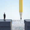 Businessman standing on cliff edge facing gap, with giant pencil drawing bridge to opposite platform above cityscape