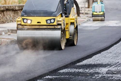 Yellow road roller compactor with headlights compacting asphalt on a construction site with additional equipment in background