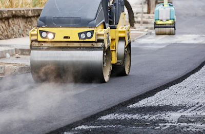 Yellow road roller compactor with headlights compacting asphalt on a construction site with additional equipment in background