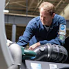 Technician in blue uniform servicing vehicle engine components in an industrial workshop setting