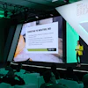 Speaker Christine Yu Moutier, MD presenting at Ground Breakers conference with green-lit stage and projection screen displaying credentials.
