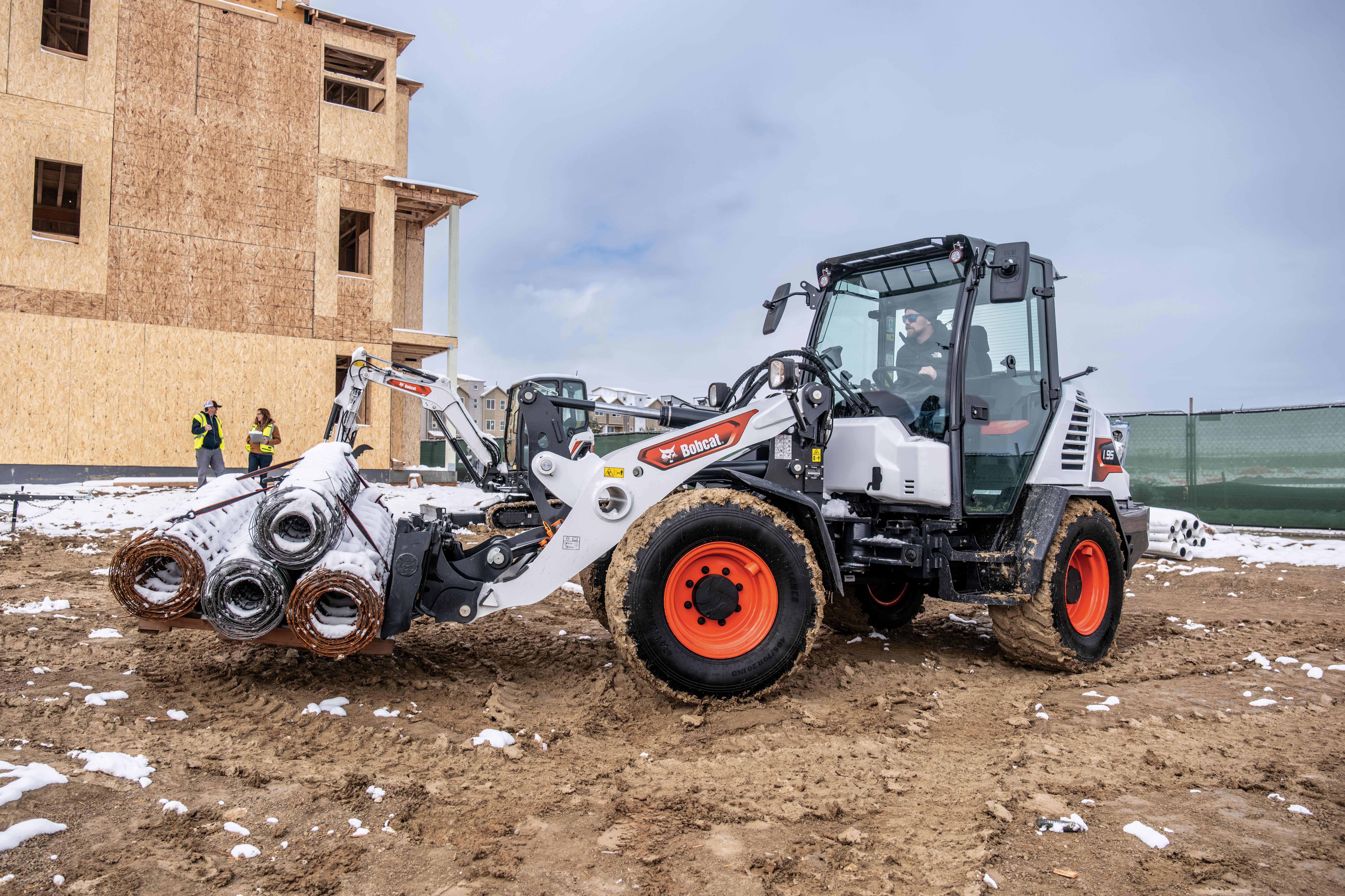 Bobcat telehandler loading metal pipes at construction site with partially built structure and workers