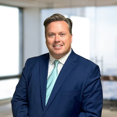 Professional headshot of a smiling man in navy blazer and light blue shirt in modern office setting