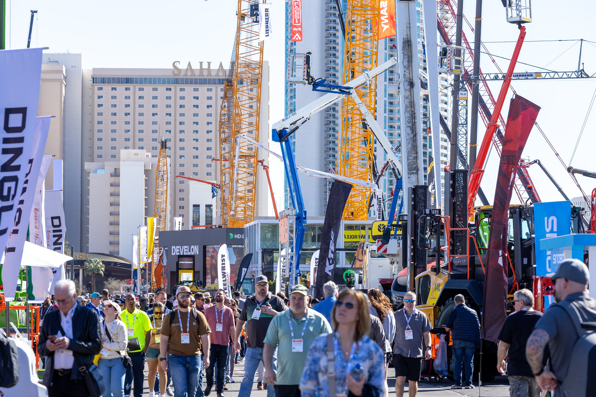 Crowded construction industry trade show with yellow and blue cranes, high-rise buildings, and attendees wearing safety gear and badges.