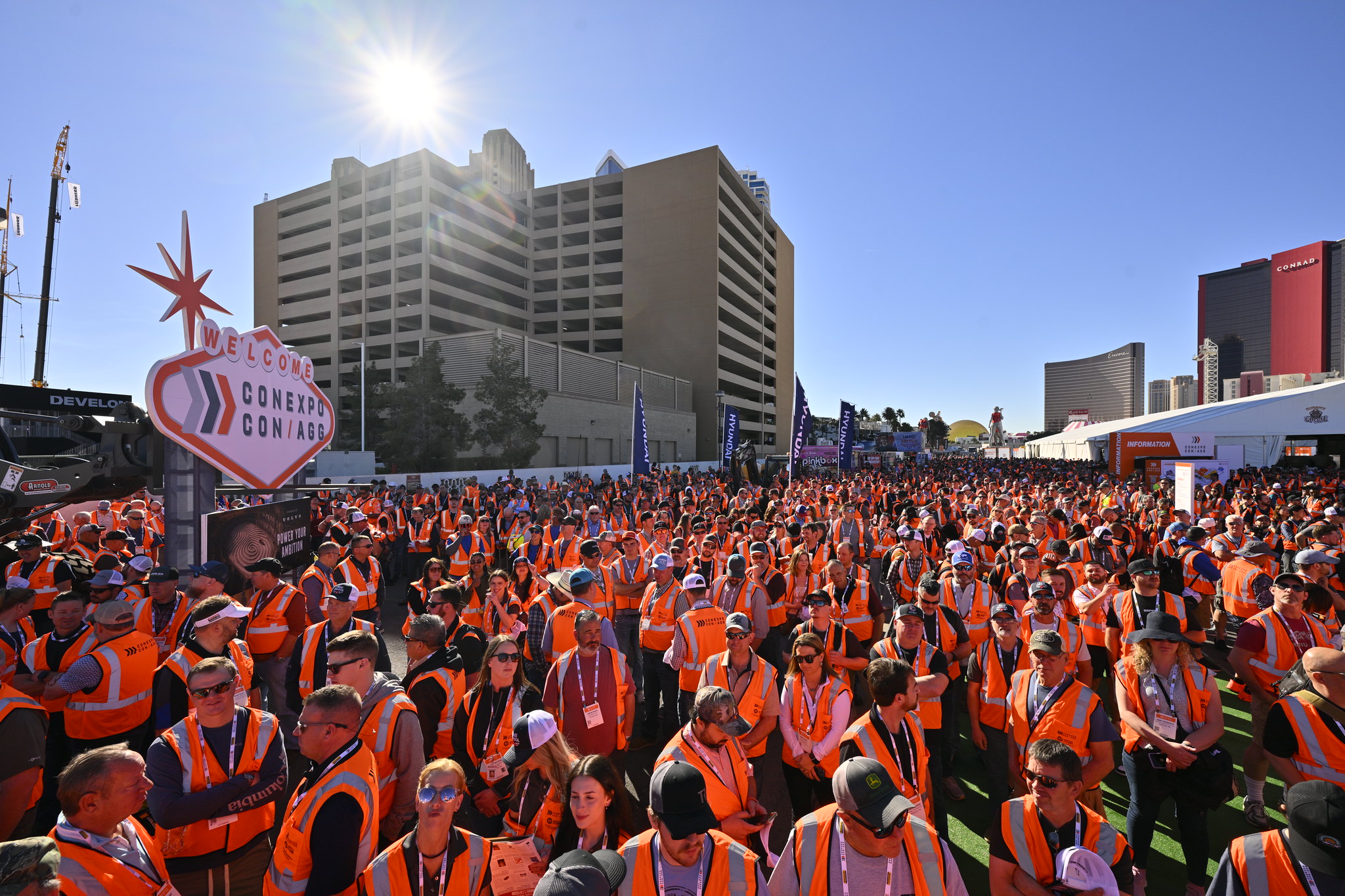 Thousands of people in orange vests gather at outdoor event with high-rise buildings and heart-shaped confetti cannon sign visible.