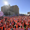 Thousands of people in orange vests gather at outdoor event with high-rise buildings and heart-shaped confetti cannon sign visible.