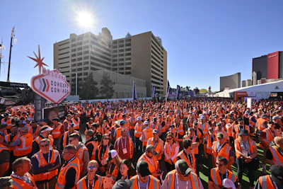 Thousands of people in orange vests gather at outdoor event with high-rise buildings and heart-shaped confetti cannon sign visible.