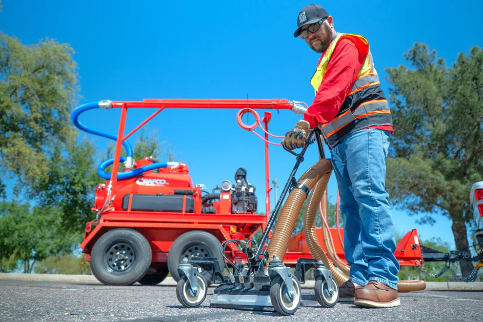 Worker in safety gear operating red industrial spray equipment on sunny day with trees in background