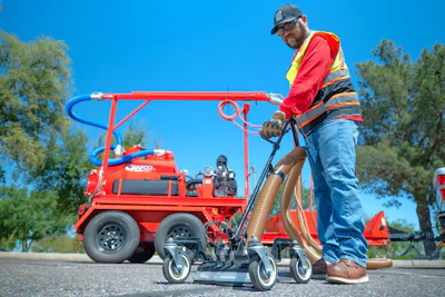 Worker in safety gear operating red industrial spray equipment on sunny day with trees in background