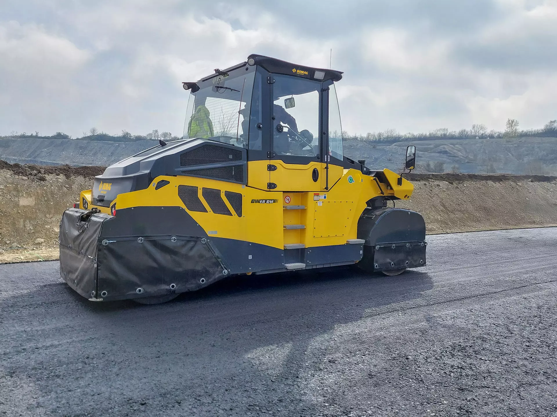 Yellow and dark blue BOMAG asphalt compactor operating on a construction site with gravel pit in background