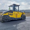 Yellow and dark blue BOMAG asphalt compactor operating on a construction site with gravel pit in background