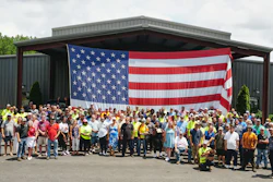 Large group of workers in safety vests and casual attire standing together in front of an industrial building with an American flag