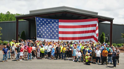 Large group of workers in safety vests and casual attire standing together in front of an industrial building with an American flag