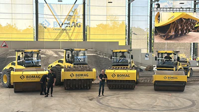 Four yellow and black BOMAG road compactors displayed in a row at an industrial facility with company signage in background