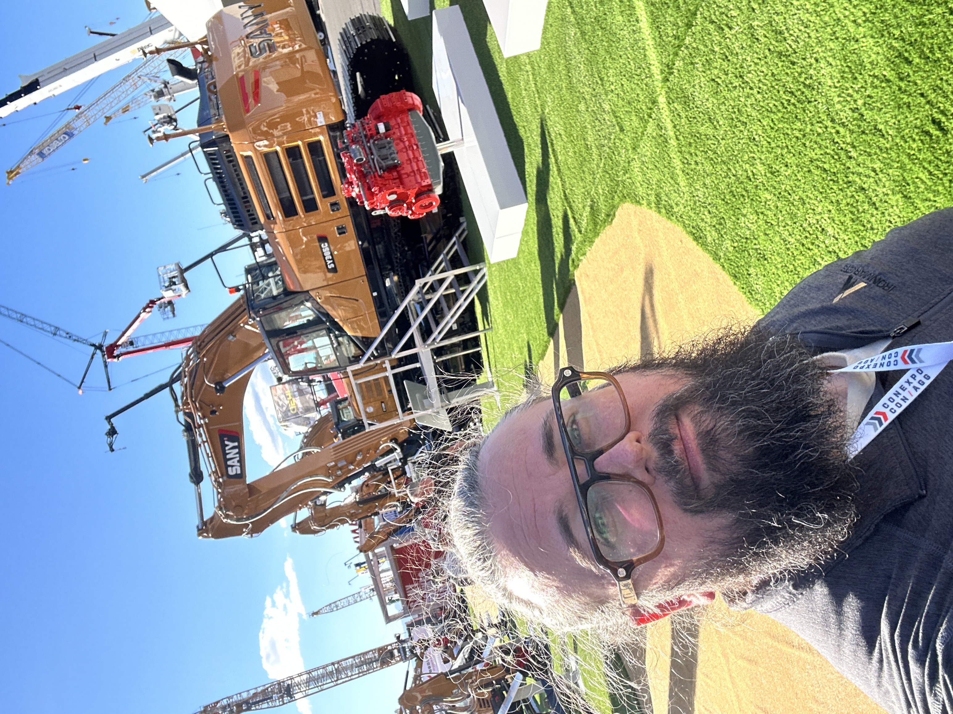 Man with beard and glasses at construction equipment trade show with SANY excavator and machinery in background