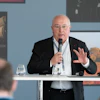 Senior professional speaking at a conference podium with microphone and water glasses
