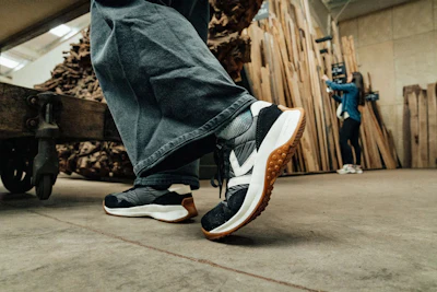 Person wearing black and white sneakers with brown sole in a workshop setting with tools and wood materials