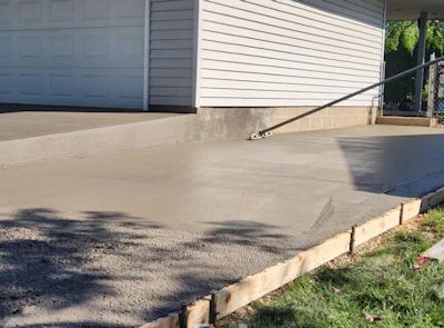 Newly poured concrete driveway with wooden forms and white garage doors visible
