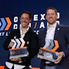 Two men in business attire holding awards at a CXGA conference event with branded backdrop
