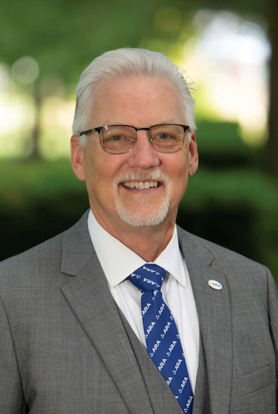Professional headshot of a man in gray suit and blue tie with glasses, smiling against a green blurred background