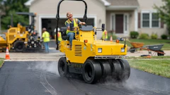 Construction worker operating a yellow SAKAI road roller compactor on a residential driveway during paving work