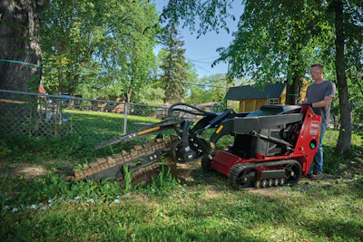 Man operating red mini skid steer loader with auger attachment in residential yard surrounded by trees