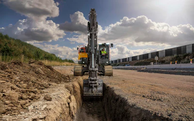 Excavator digging trench at construction site with yellow truck and industrial buildings in background under cloudy sky