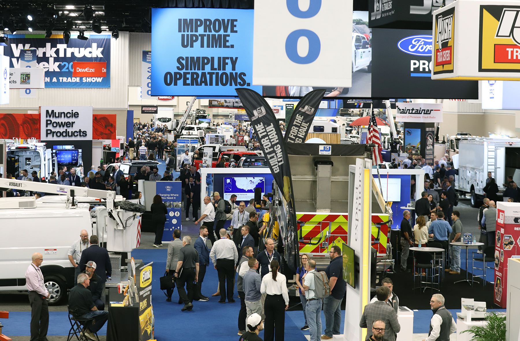 Busy trade show floor with multiple vendor booths, overhead signage, and crowds of attendees at an industrial or automotive exhibition.