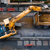 Yellow excavator loading construction debris into truck on urban street with benches and buildings