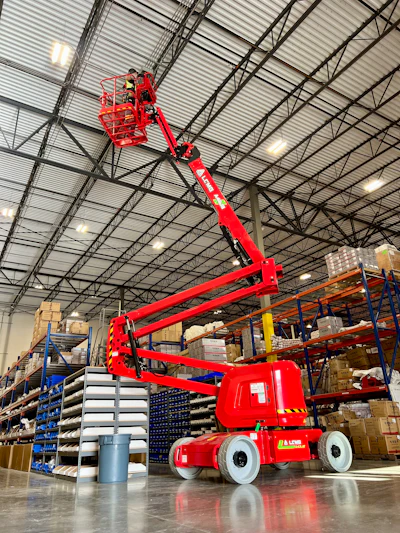 Red articulating boom lift in warehouse with blue and orange storage racks and industrial ceiling