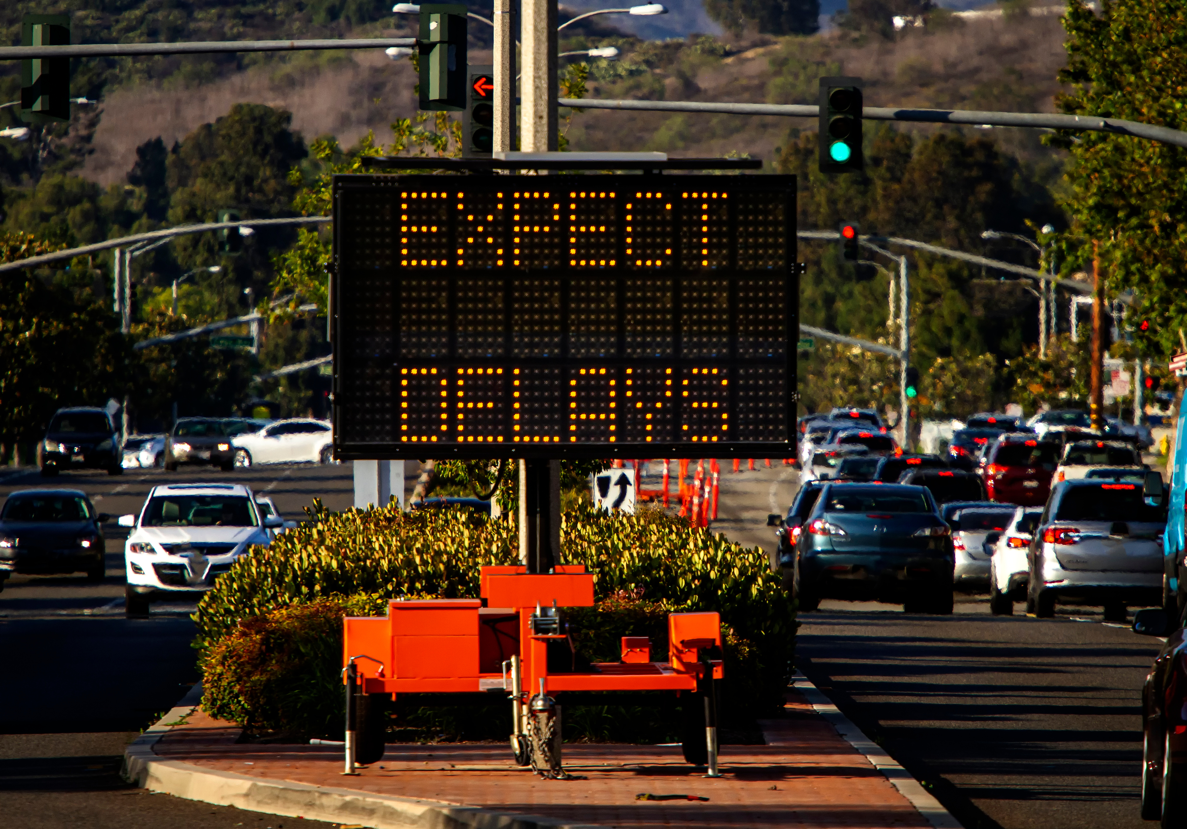 Digital traffic sign displaying 'EXPECT DELAYS' in amber text above a parking lot with vehicles and landscaping.