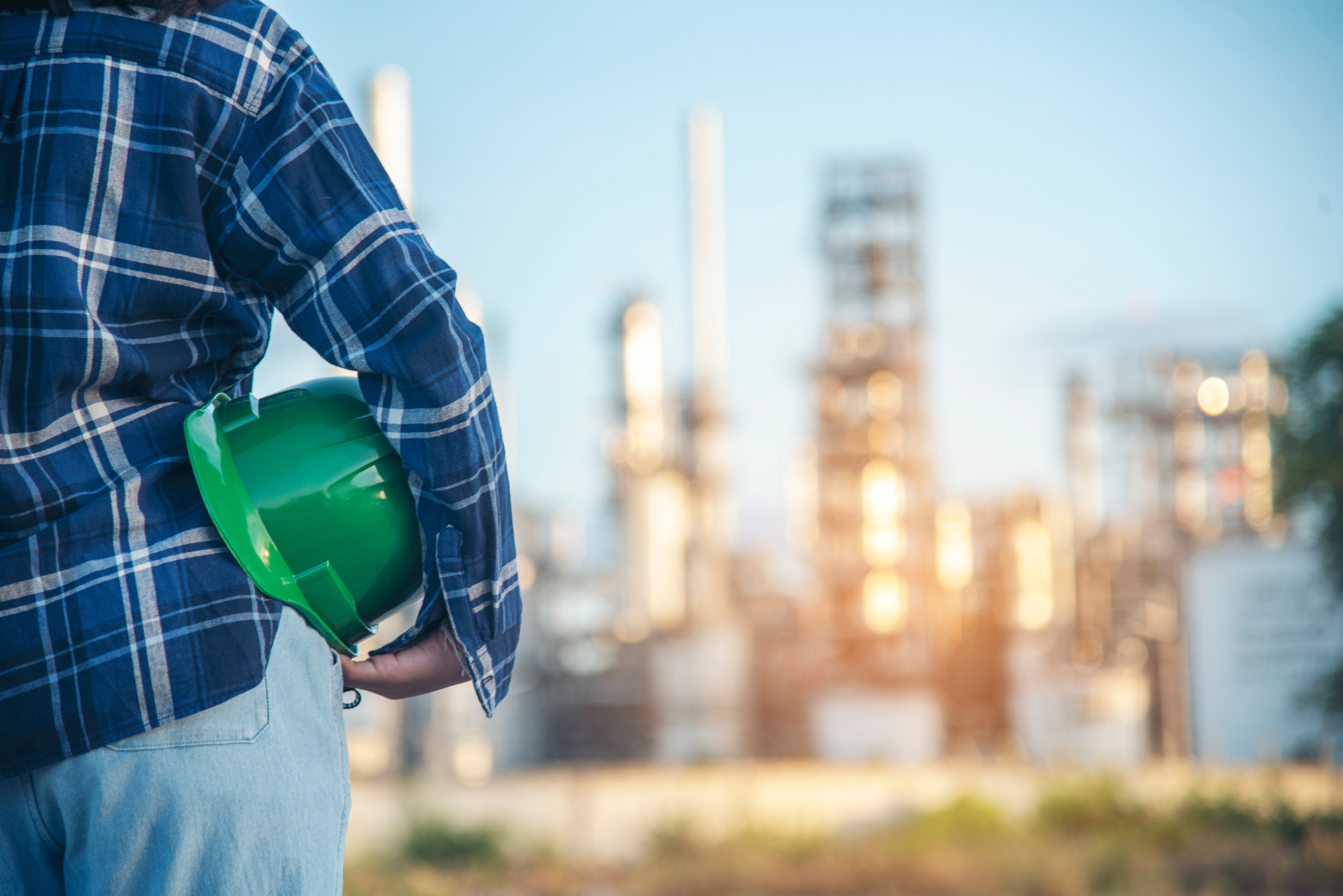 Construction worker in blue plaid shirt holding green hard hat with industrial refinery in background